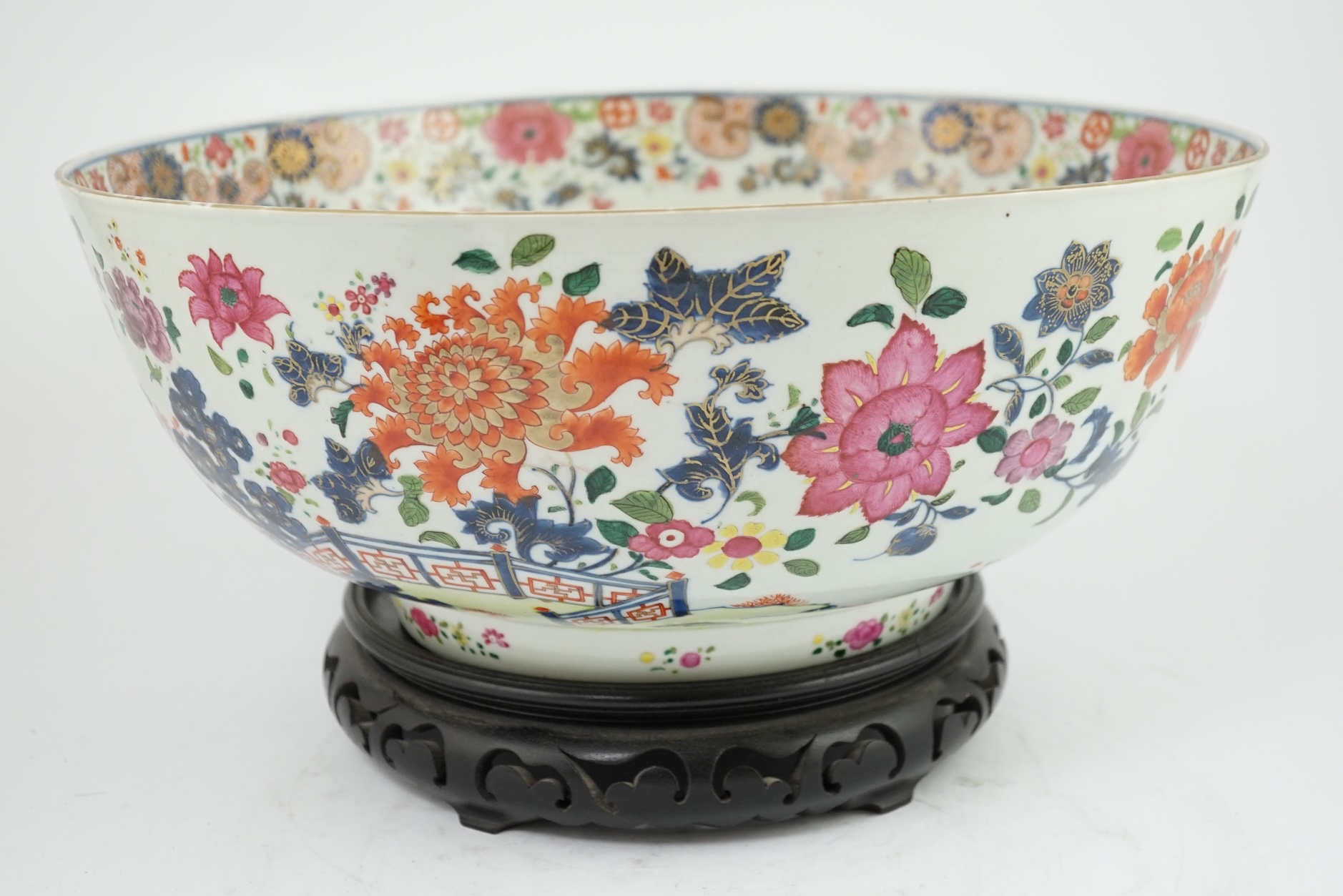 A large Chinese famille rose punch bowl, Qianlong period, the exterior painted with flowers in a fenced garden, the interior with a flower bouquet within a garland of flowers, 40.5cm diameter, wood stand. Condition - som